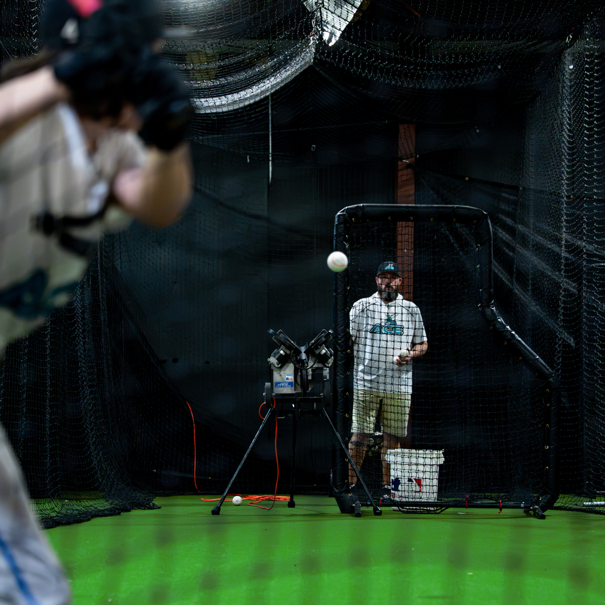 A man stands inside a batting cage wearing a white t-shirt and green shorts, holding a Velomax baseball near a Jr Hack Attack pitching machine with Timing Chute. Another person is blurred in the foreground on the left, and the background shows black netting and a green turf floor.