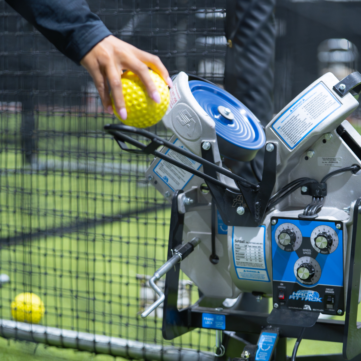 A close-up action shot of a person's hand tossing a softball into a extended chute attached to a Jr Hack Attack Softball Machine.