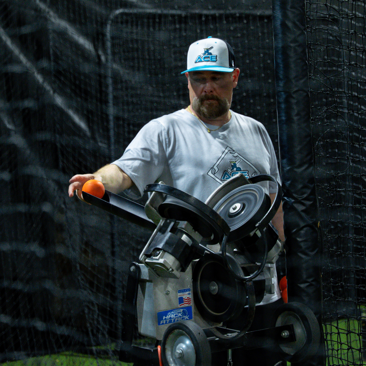 A bearded man in a white and blue baseball cap stands behind a Jr Hack Attack Pitching  Machine with timing chute, holding an orange velomax