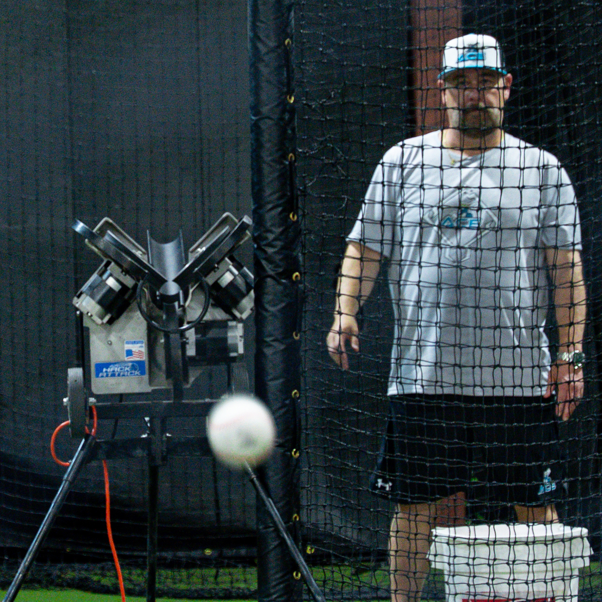 A man wearing a gray t-shirt, black shorts, and a white cap stands behind a batting cage net, waiting for a Velomax baseball in flight that is blurred in the foreground. Jr Hack Attack Pitching Machine with Timing Chute  is visible on the left.