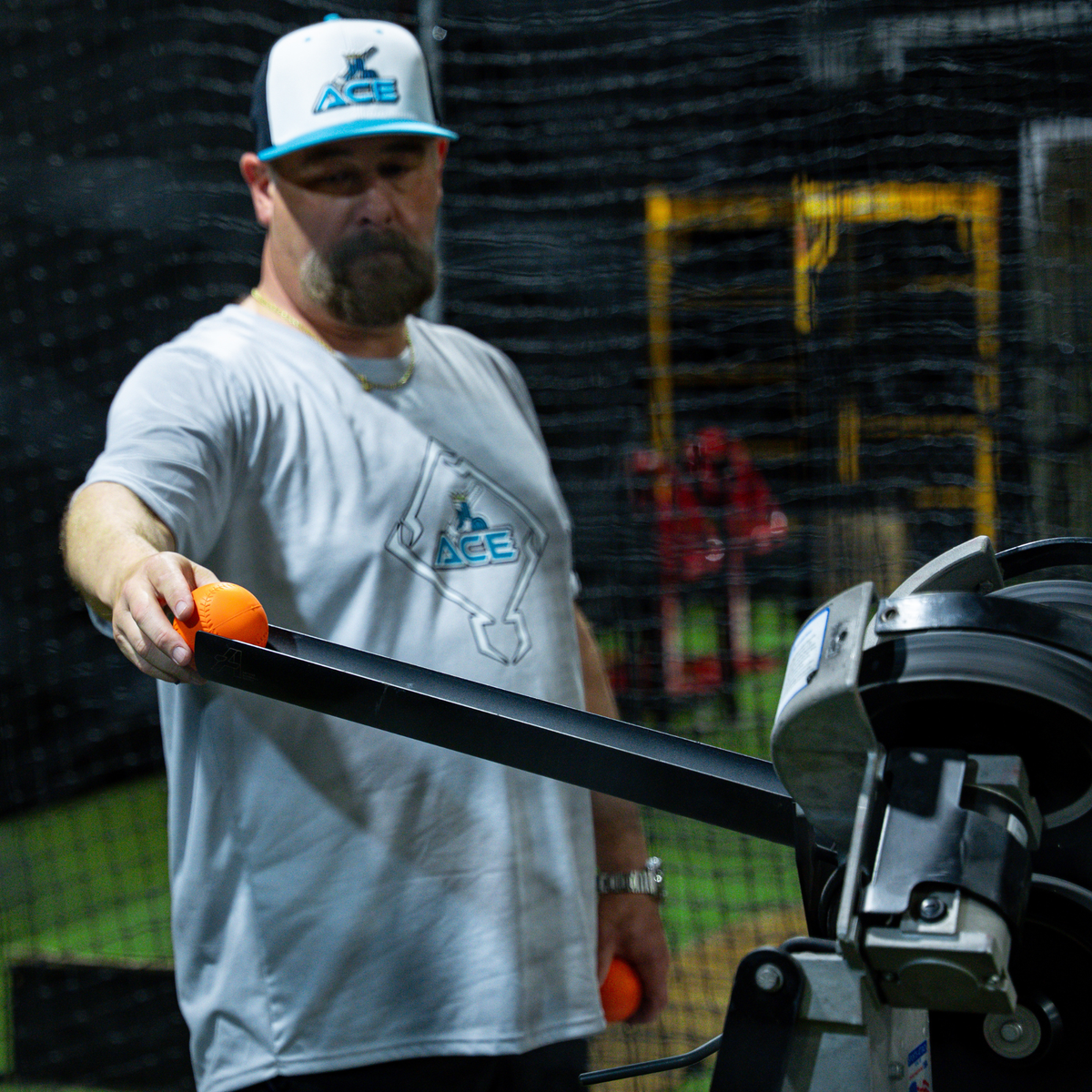 A bearded man wearing a white and blue Ace brand cap and matching t-shirt stands in a batting cage, holding an orange velomax next to the Jr Hack Attack Pitching Machine with Timing Chute