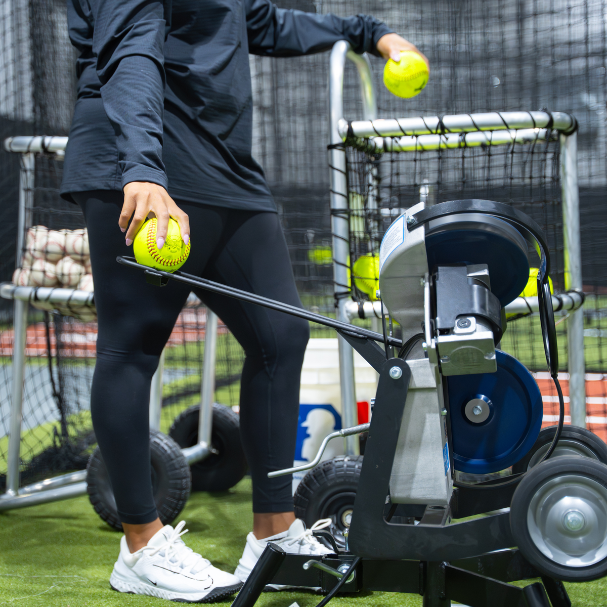 A person in black athletic wear stands at a Jr Hack Attack Pitching Machine with Timing Chute for Softball, holding yellow softballs in a practice field setting with batting cages and turf visible.