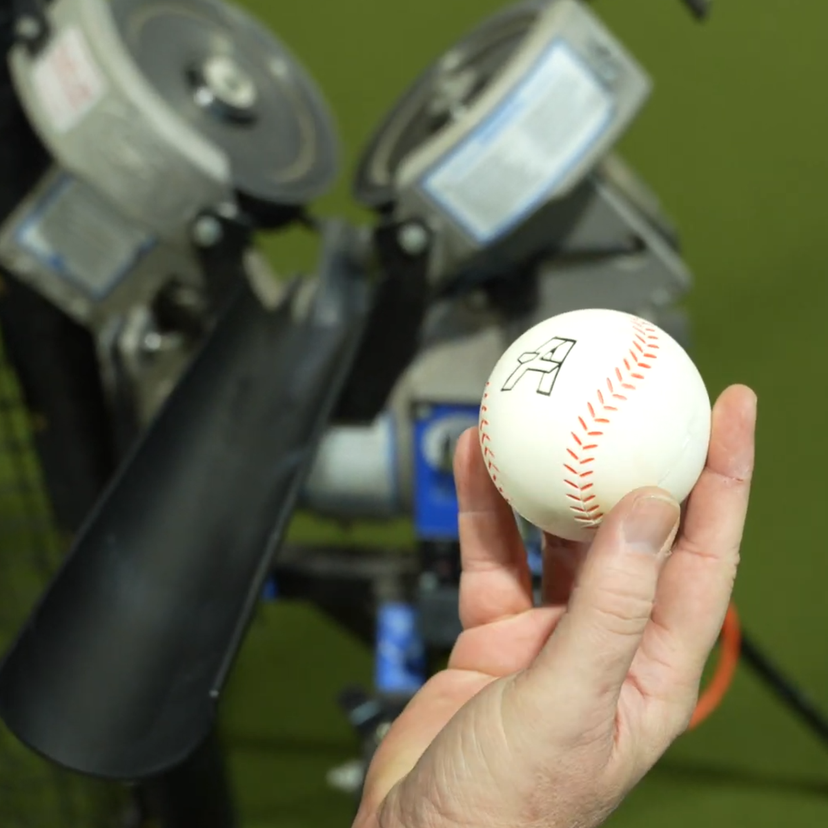 A hand holding a white Velomax, with a blurred baseball training machine in the background.